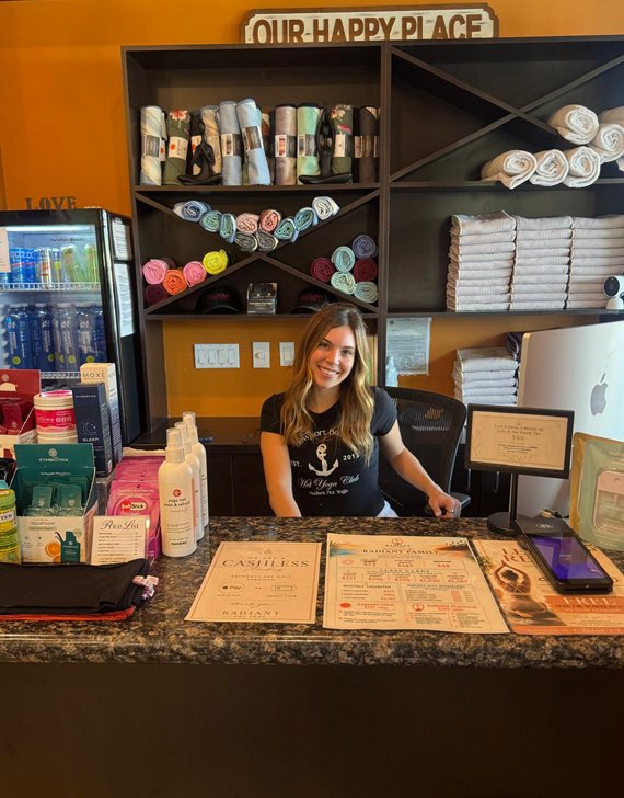 Smiling receptionist sits at a dark marble counter in a spa shelves of rolled and folded towels behind her